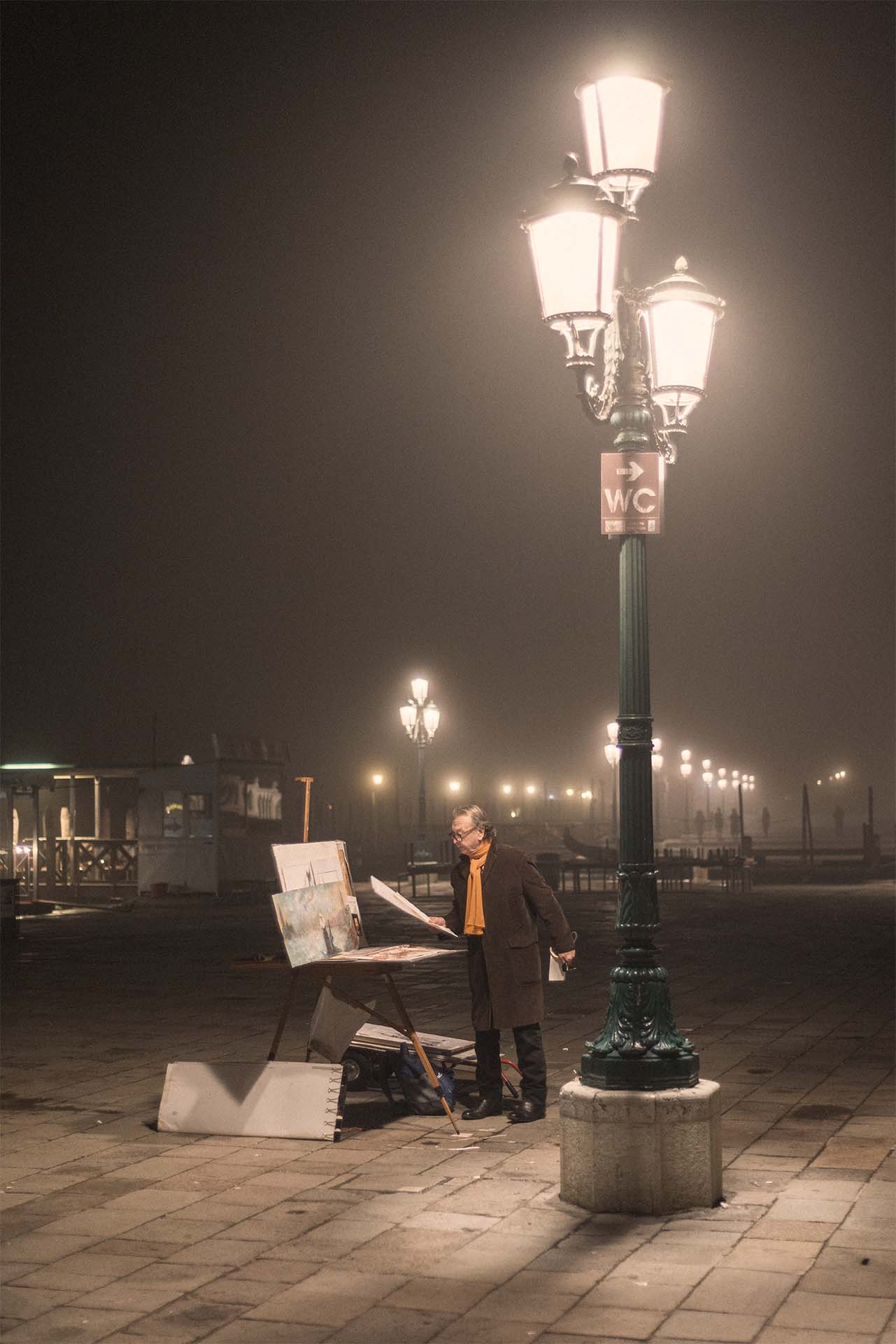 An image of a painter looking at a painting in his hand next to a street lamp in San Marco Square of Venice in a freezing foggy night.