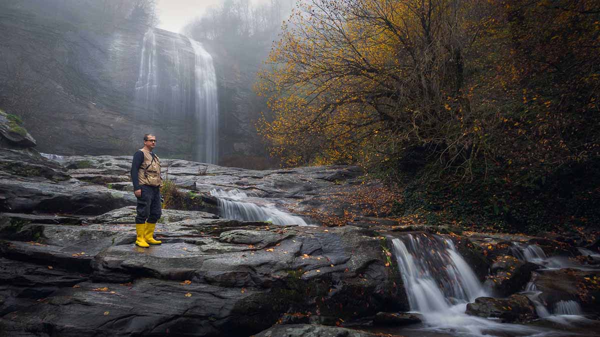 A man standing over some rocks next to a waterfall in an autumn setting with orange leaves on the ground.